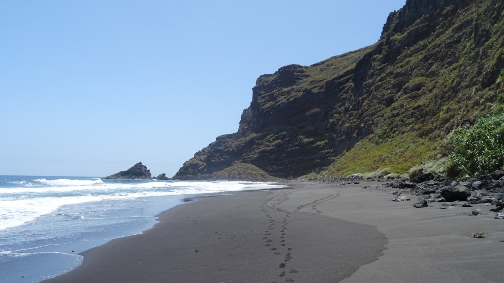 Une plage de sable noir avec des empreintes de pas, bordée par des falaises verdoyantes sous un ciel bleu clair.