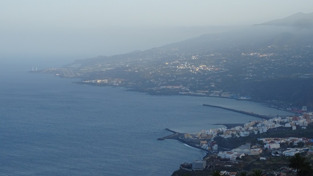 Vue panoramique du littoral avec des bâtiments colorés, l'océan et les montagnes en arrière-plan, sous une lumière brumeuse.