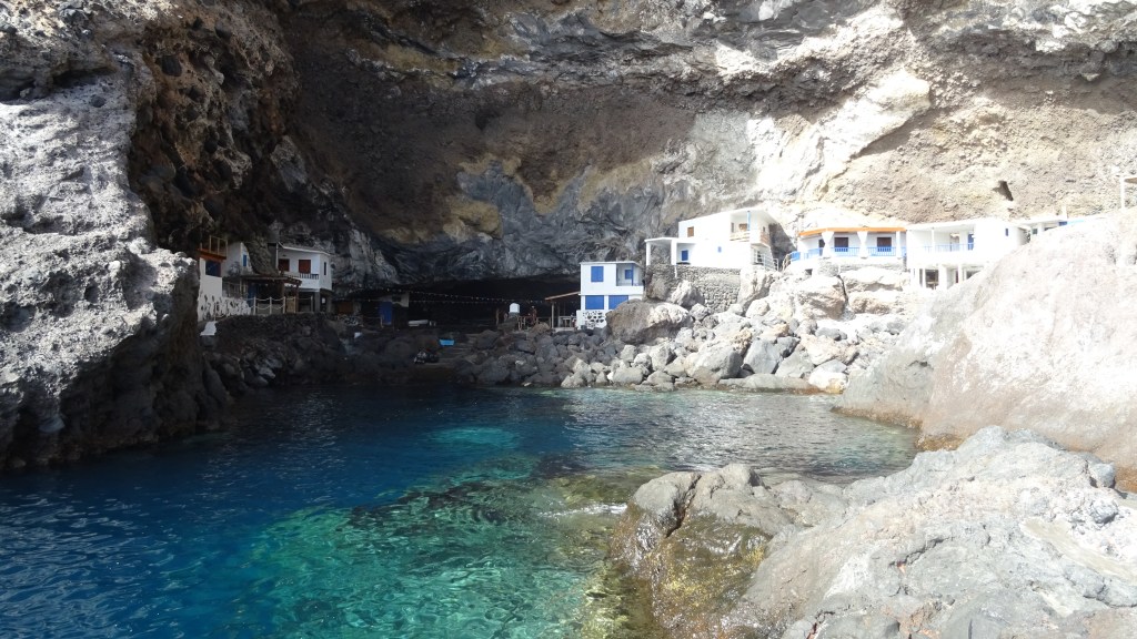 Vue d'une grotte marine avec des maisons blanches et bleues au bord de l'eau, entourées de rochers et d'une eau turquoise.