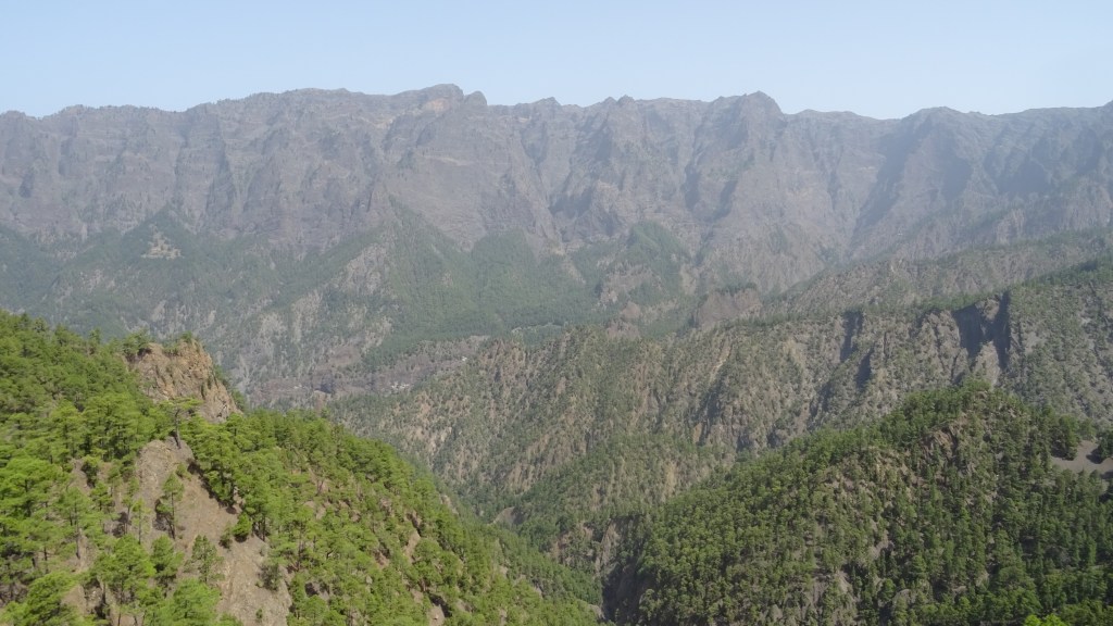Vue panoramique des montagnes et des forêts verdoyantes, avec des pentes escarpées et des sommets rocheux sous un ciel clair.
